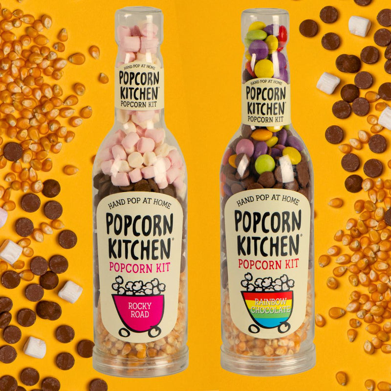 2 bottles of pop at home popcorn kits on an orange background with popcorn kernels, chocloate buttons and marshmallows scattered around them