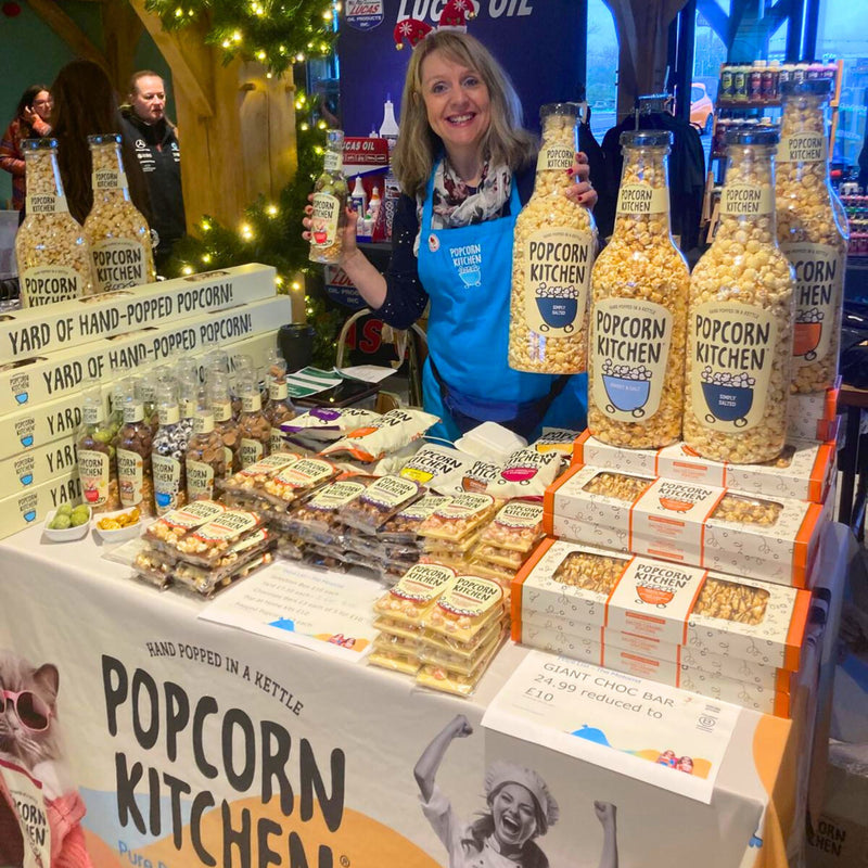 woman standing behind a table filled with popcorn bottles and popcorn chocolate bars