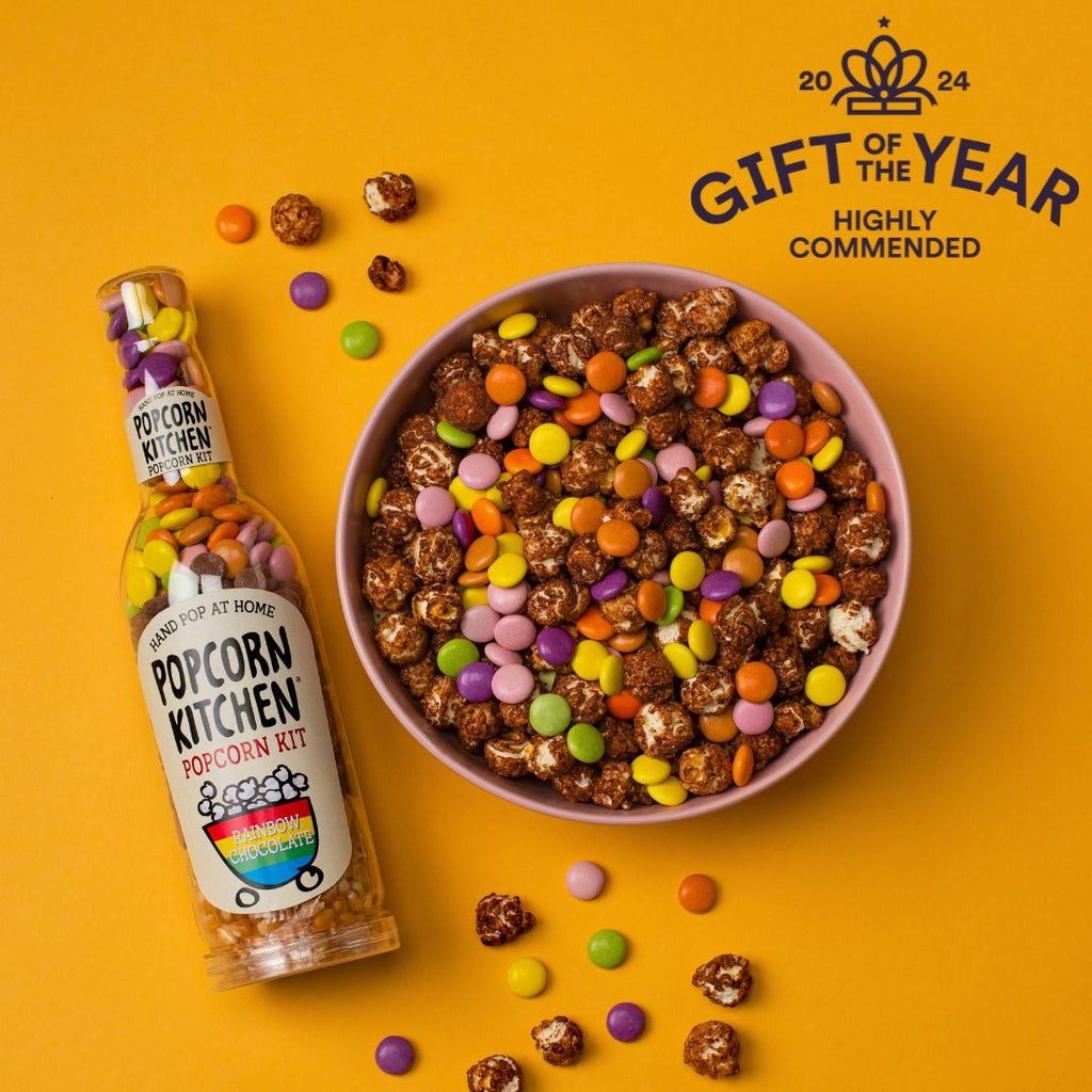 Pop At Home Rainbow Chocolate Bottle next to a bowl containing chocolate coated popcorn and rainbow chocolates. Gift of the year highly commended 2024 logo is showing