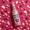 A bottle of white chocolate and raspberry popcorn lying flat on a pink background with popcorn, raspberries and white chocolate pieces scattered around it.