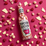 A bottle of white chocolate and raspberry popcorn lying flat on a pink background with popcorn, raspberries and white chocolate pieces scattered around it.